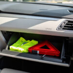 Quels équipements obligatoires et indispensables garder dans sa voiture ? Habitacle moderne d’une voiture avec un gilet fluorescent rangé dans la boîte à gants ouverte et un triangle de présignalisation dans son étui, éclairé par une lumière naturelle.