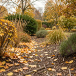 Jardin d’automne réaliste avec des feuilles mortes dorées autour d’arbustes et de plantes vivaces, sur un sol recouvert de paillage naturel, baigné d’une lumière douce.